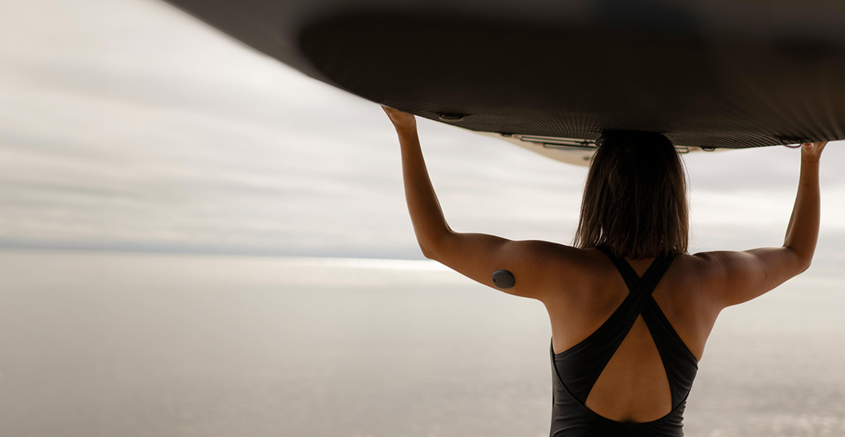A person carrying a surfboard overhead stands on a beach, with a calm ocean and overcast sky in the background.